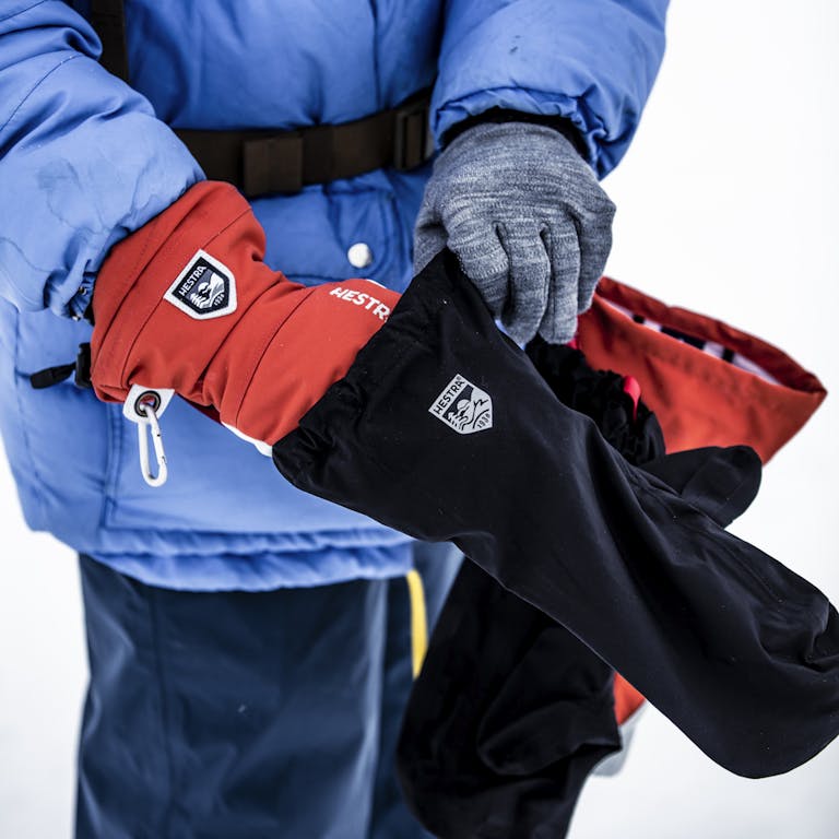 _E1A8702 (1) En person tar på sig en svart skalhandske över en röd skidhandske och grå linerhandske i snöigt landskap