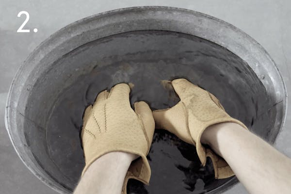 steg 2 dress En handskmakare med bruna skinnhandskar på sig doppar händerna i en balja med vatten i ett handskmakeri