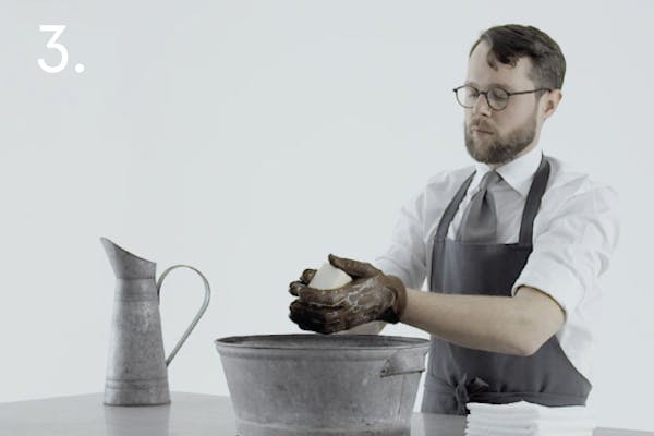 steg 3 dress En handskmakare med blöta skinnhandskar på sig tvålar in handskarna över en balja i ett handskmakeri