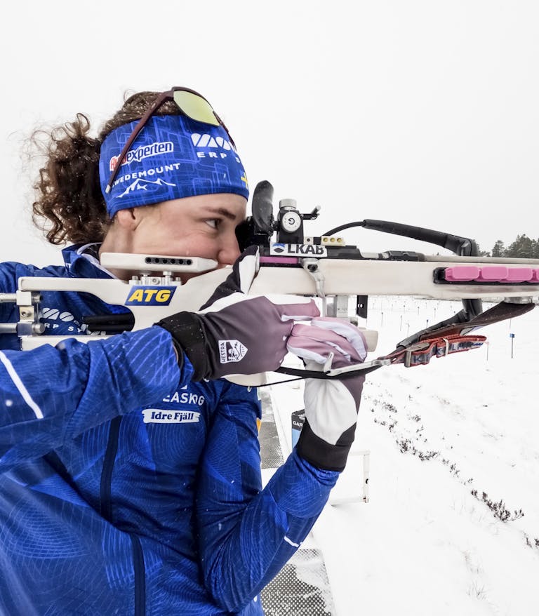 DCIM100GOPROGOPR1256.JPG En kvinna i blå skidkläder siktar med ett gevär under ett skidskytte evenemang i snöiga förhållanden