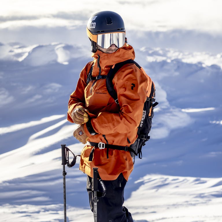 _D5A9289-Förbättrat-BR En man i orange skidjacka och hjälm spänner sin jacka på en snötäckt plats medan han bär skidutrustning