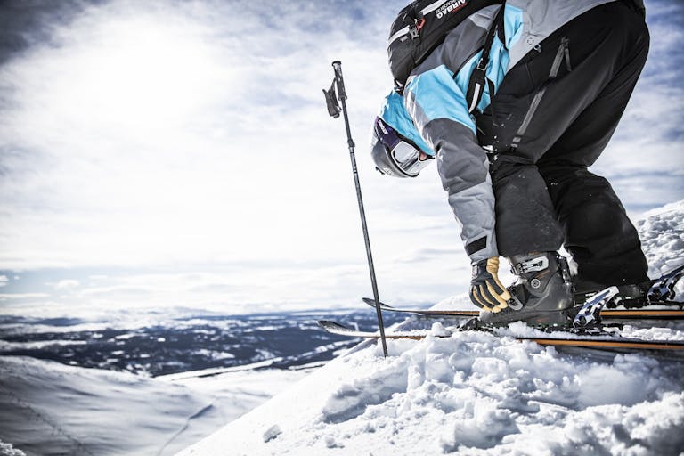 _E1A8059 En man justerar sina skidor på en snötäckt bergstopp med skidhandskar i snöiga förhållanden