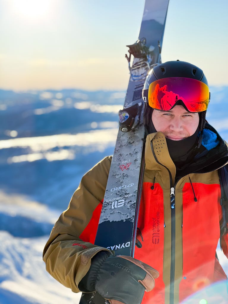 IMG_5335 En skidåkare i tvåfärgad skidjacka bär sina skidor på axeln under en solig dag i fjällmiljö