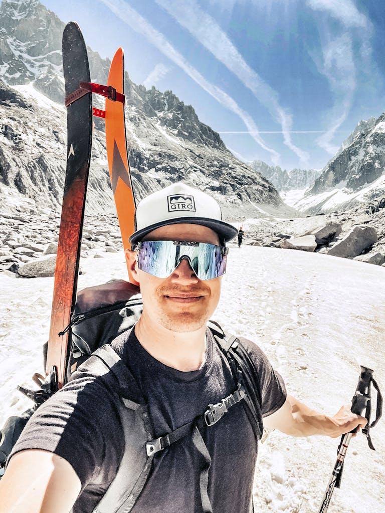 IMG_8177 En man med skidor på ryggen tar en selfie i soligt fjällandskap med snö och berg i bakgrunden