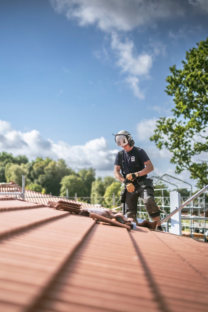 A man in workwear standing on a roof with blue sky and green trees in the background.