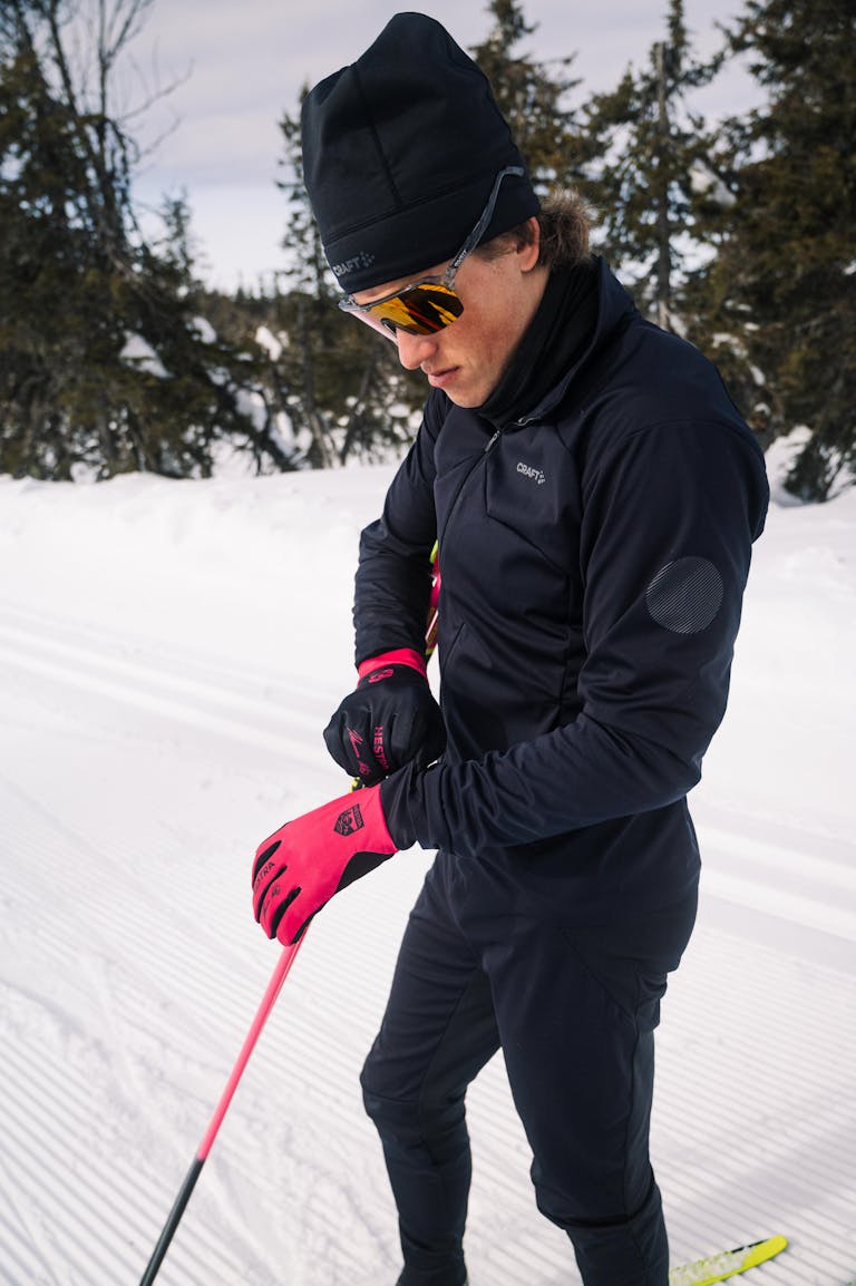 A man adjusts his gloves while standing on snow-covered tracks in ski clothing, preparing for cross-country skiing