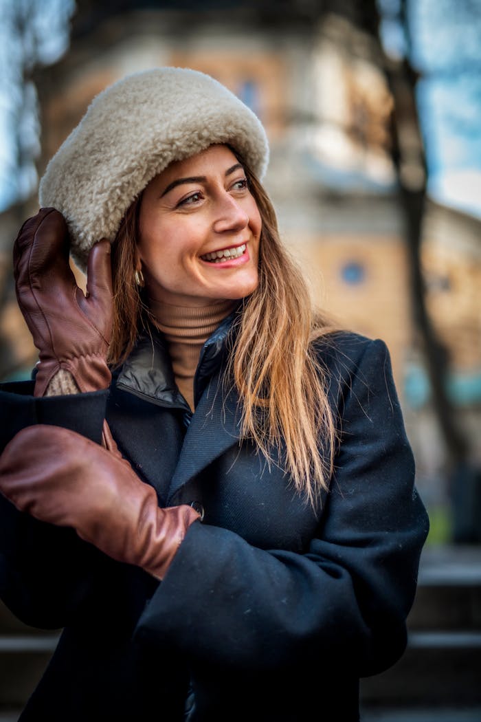 A smiling woman wears brown leather gloves, a dark coat, and a warm hat on a cold day in the city
