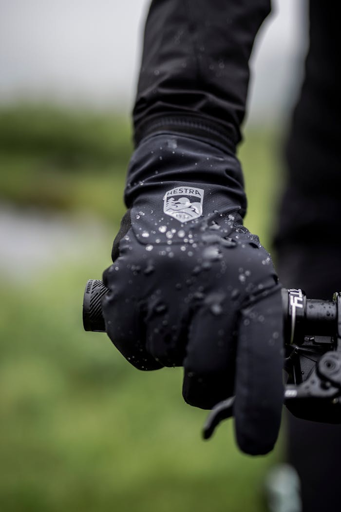 A cyclist in cycling gloves grips the handlebars during a rainy ride through nature, with raindrops on the gloves