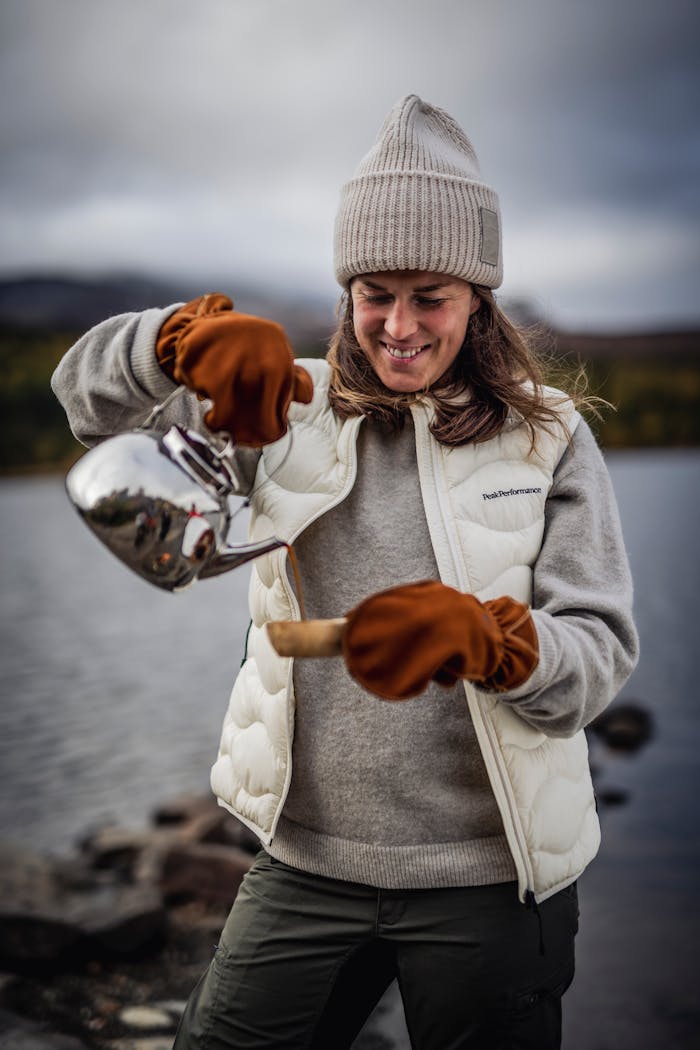 A smiling woman in autumn clothing pours coffee from a thermos by the lake, surrounded by calm nature and muted colors