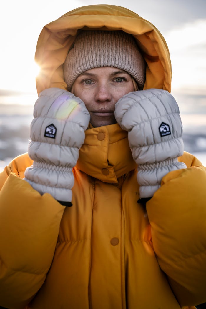 A person in winter clothing stands in a winter landscape with the sun behind them, hands to their face to stay warm